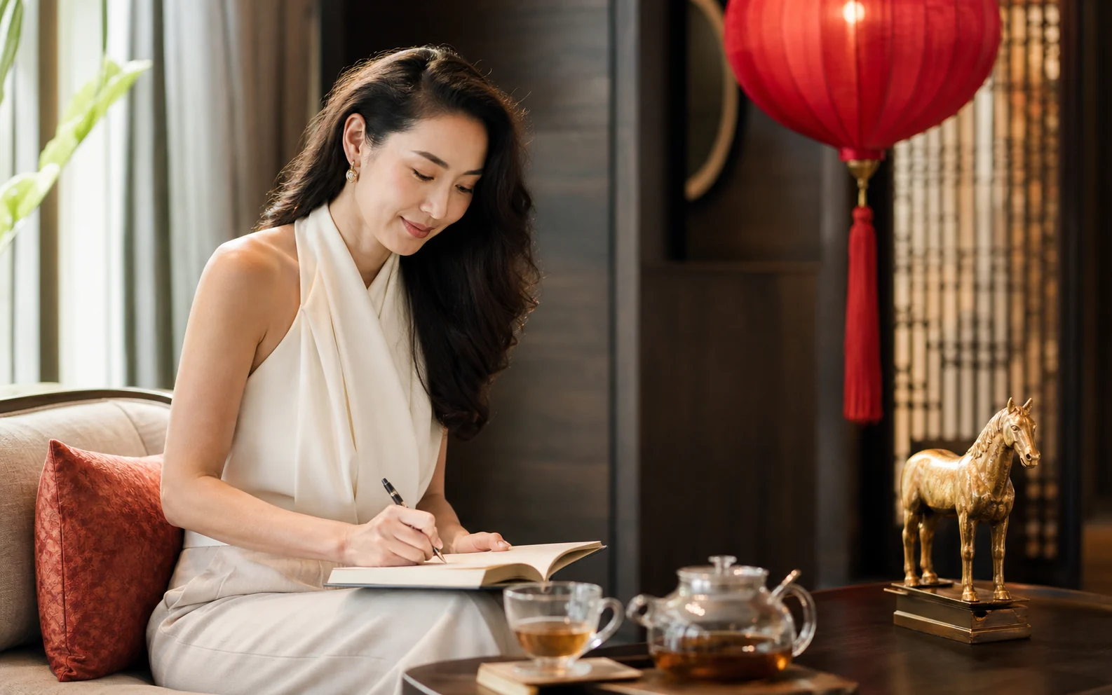 Thai-Chinese woman journaling beside herbal tea, a red lantern and a brass horse in a Bangkok wellness lounge
